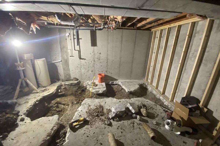 Room under renovation with broken concrete flooring, exposed soil, wall studs, pipes, tools and an orange bucket