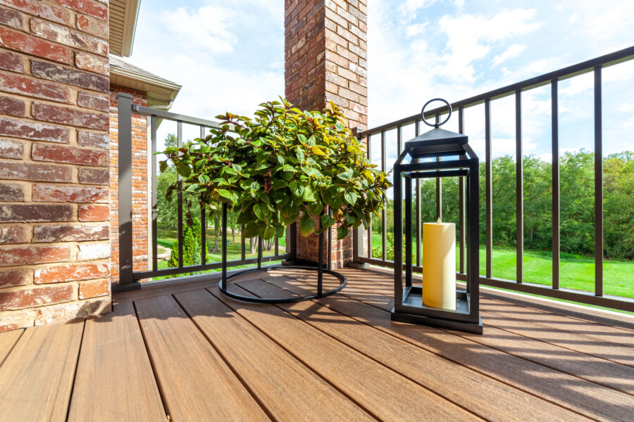 Covered porch corner with a potted plant on a black metal stand and a candle lantern, beside a brick column and railing