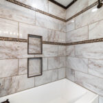 White bathtub surrounded by marble-patterned tile, mosaic trim, two recessed niches and dark fixtures