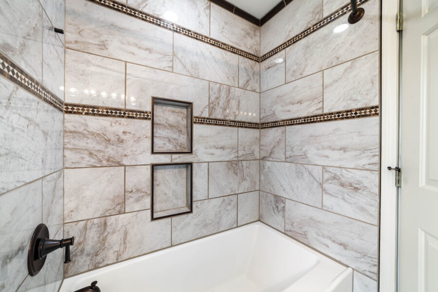 White bathtub surrounded by marble-patterned tile, mosaic trim, two recessed niches and dark fixtures