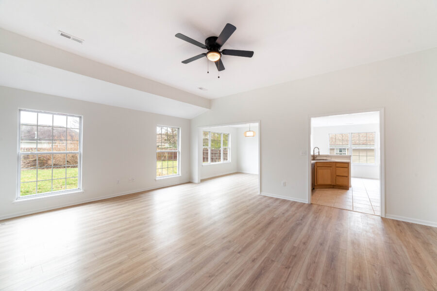 Empty living room with gray walls, wood-look plank flooring, a ceiling fan, and an opening to a kitchen