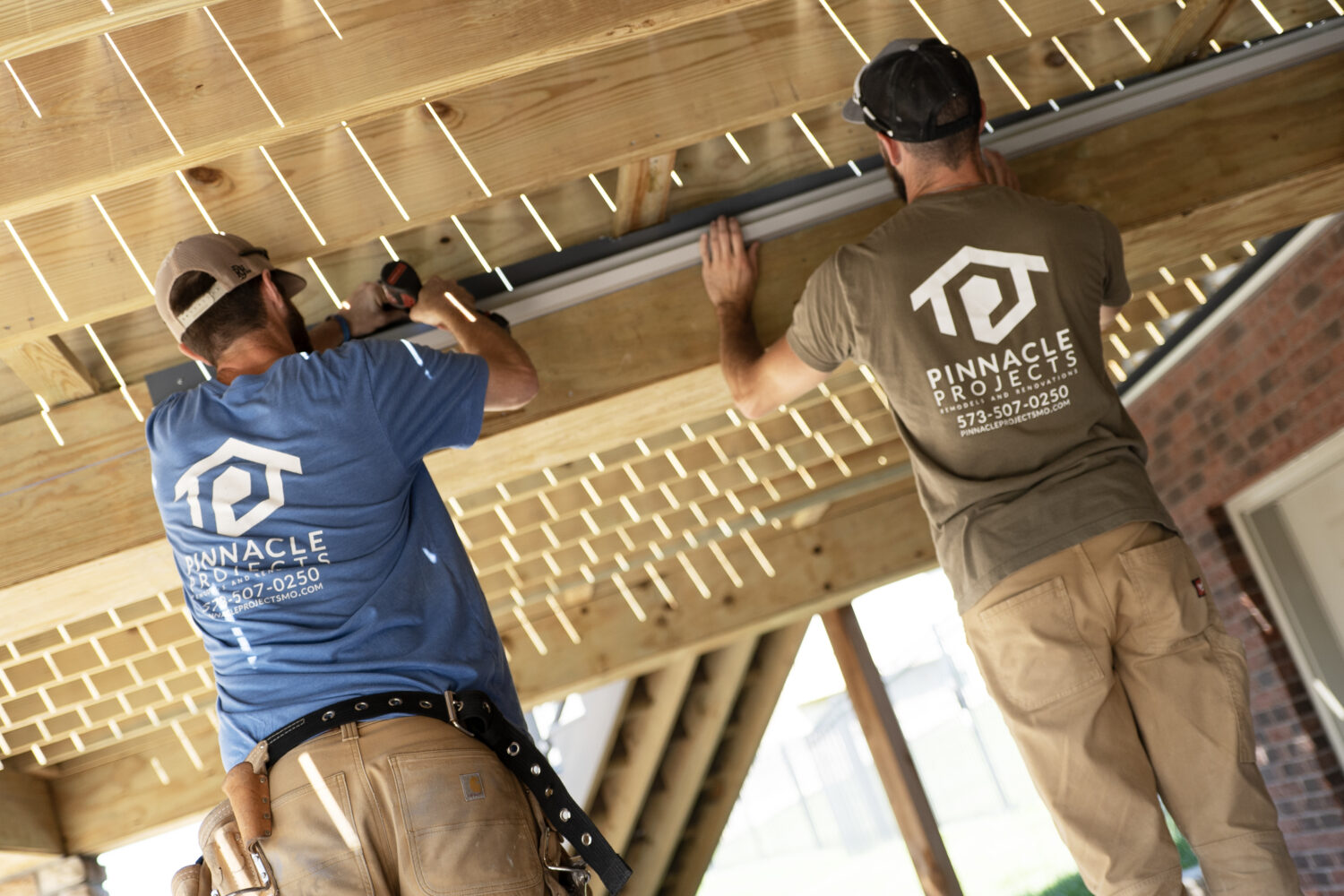 Two workers using a power tool on a gray metal rail beneath exposed wood beams and boards, next to a brick wall