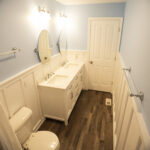 Bathroom with a white double-sink vanity, two oval mirrors, towel bars, a closed door and wood-look flooring