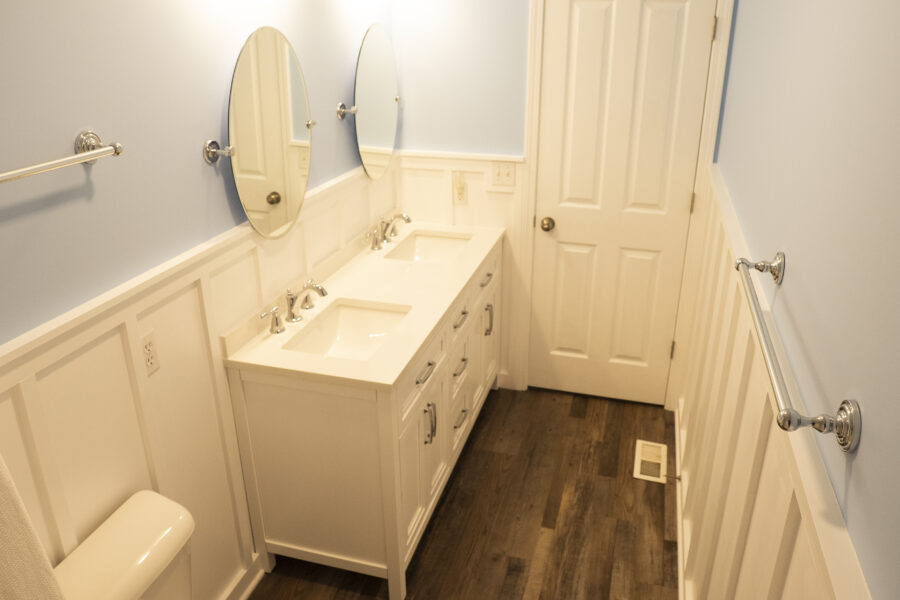 Bathroom with a white double-sink vanity, two oval mirrors, towel bars, a closed door and wood-look flooring