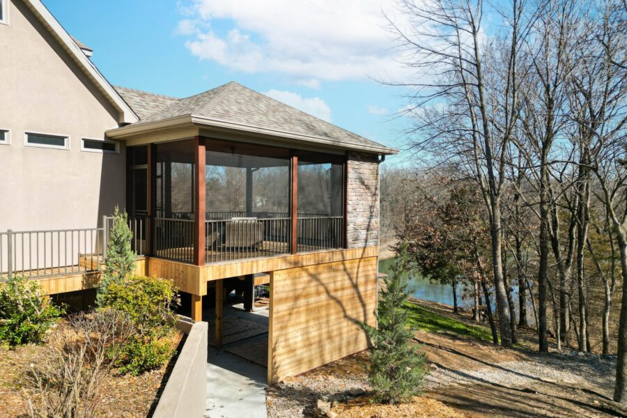 House with an elevated screened porch on wooden posts, shrubs and leafless trees overlooking water