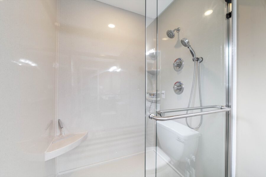 Glass-enclosed shower with beige walls, two chrome showerheads, round controls and a corner shelf
