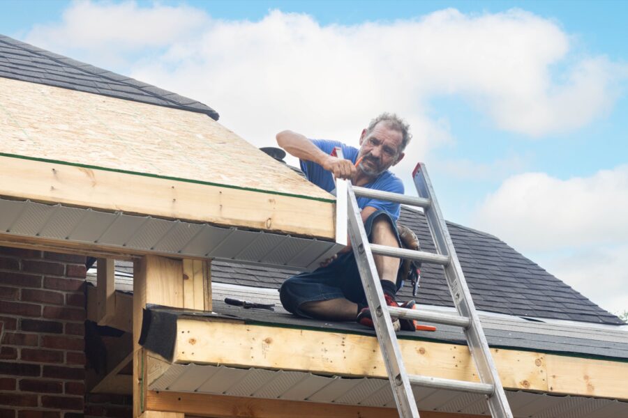 Man kneeling on a partly finished roof beside a metal ladder, working on exposed wooden trim